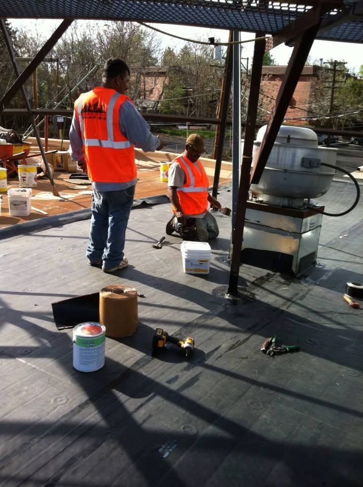 Two roofers on a flat roof in orange safety vests; the vest on the left shows 303-437-5915